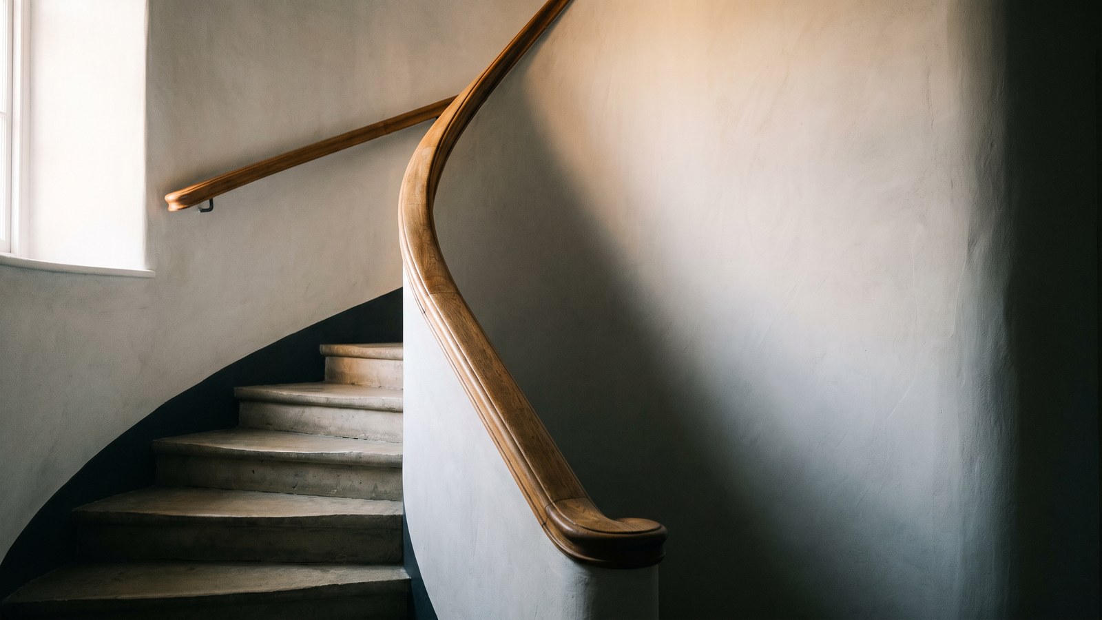 A pale limestone staircase curving down through a lime-plastered stairwell, a smooth hand-worn oak handrail following the curve of the wall and ending in a rounded newel, soft morning light from a window at the left catching the rail and the worn stone treads.