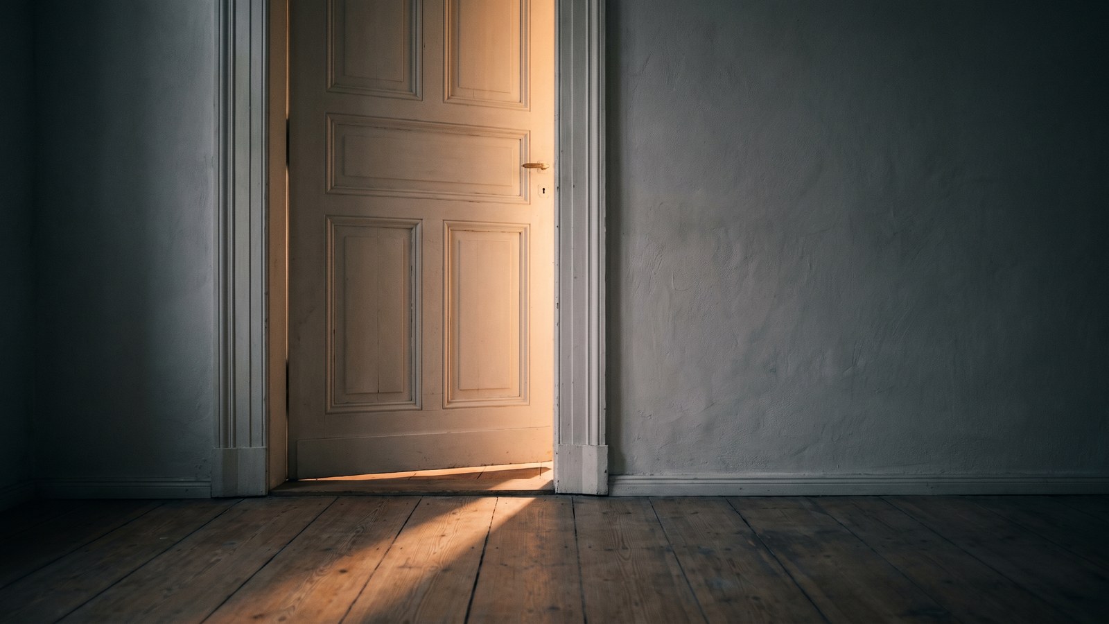 A tall panelled off-white door standing ajar in a bare room, warm morning light spilling through the gap and falling in a single shaft across wide pine floorboards, a plaster wall in soft shadow beside it.